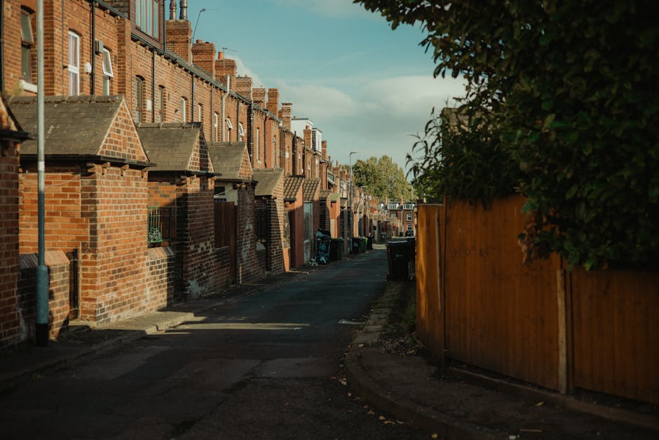 A narrow residential alleyway lined with red-brick terraced houses featuring small stepped front gardens and pitched tiled roofs. The pavement and driveway are dark asphalt, with some sunlight casting